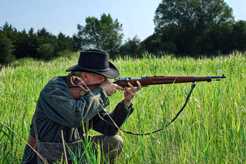 Hunter crouches in the grass and aims his rifle