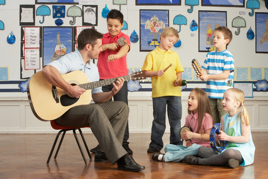 Male Teacher Playing Guitar With Pupils Having Music Lesson In C