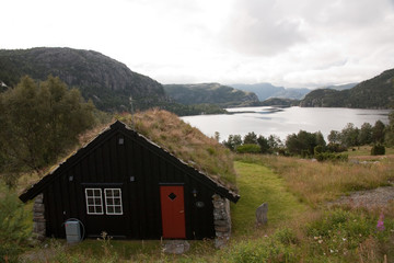 Fototapeta na wymiar a norwegian little and typical wooden house with a red door and roof full of grass near a lake between the mountains in a cloudy summer day