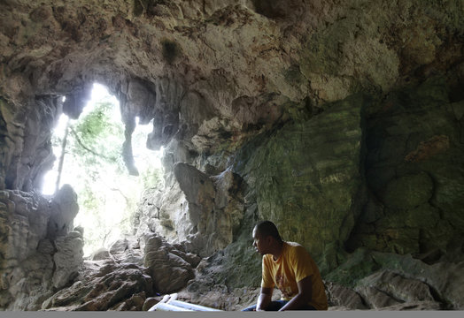 A man sits quietly inside a dimly lit cave, surrounded by natural rock formations and rugged textures. The scene captures a peaceful and reflective moment in nature