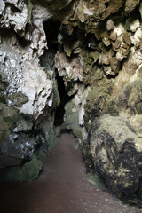 A narrow and dimly lit limestone cave featuring sharp, uneven rock formations and rugged walls. The atmosphere is mysterious and slightly eerie, making it suitable for themes such as adventure, geolog