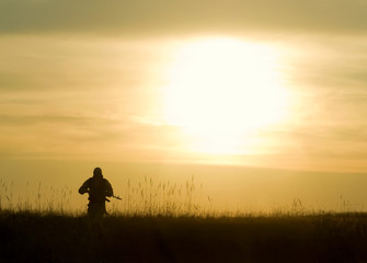 Man with rifle walking on grassy landscape during sunset