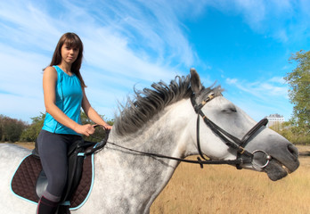 Obraz premium A young woman wearing a blue tank top rides a white horse through a field under a bright sky. The equestrian scene captures a calm moment of connection between the rider and her dappled mount.