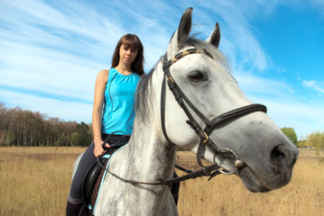 Fototapeta premium A young woman in a blue top enjoys a sunny horseback ride on a white horse through a dry field under a blue sky with clouds.