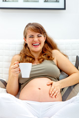 Obraz na płótnie Canvas Laughing pregnant woman sitting on sofa with cup of tea