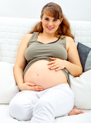 Smiling pregnant woman sitting on sofa and touching her belly.