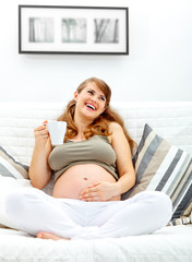 Obraz na płótnie Canvas Laughing pregnant woman relaxing on couch with cup of tea