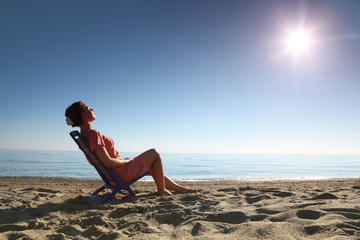Woman sits on  plastic chair on sea-shore by person to sun