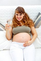 Smiling pregnant woman relaxing on sofa with glass of water.