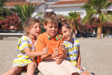 little boy and two girls sitting on beach and holding lollipops