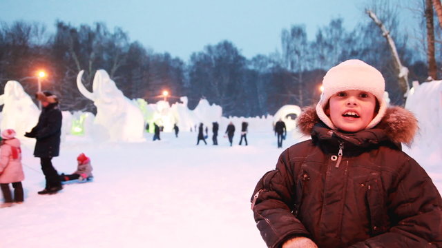 boy is telling camera on evening, behind snow sculpture