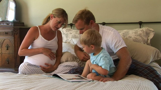 Pregnant mother, father and toddler son reading book on bed