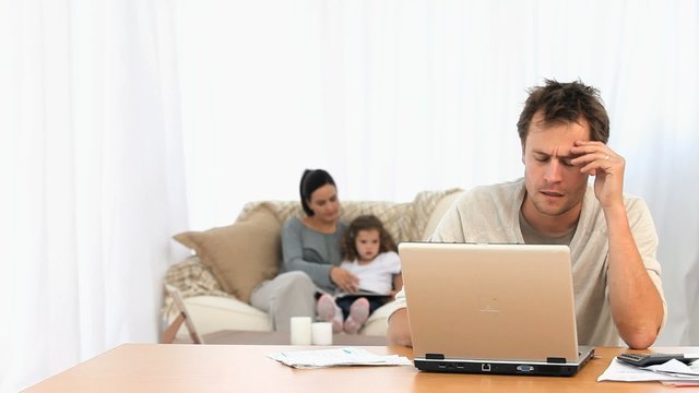 Angry man on his laptop while his family is on a sofa