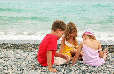 little brother and two sisters are sitting on beach and play.