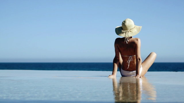 Woman in summer hat sitting in infinity pool and taking sunbath