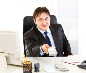 Businessman sitting at office desk and giving dollar pack.