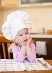 Little cook girl sitting at kitchen table waiting for meal