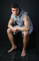 Muscle young boy sits in grey clothes on a black cube in dark