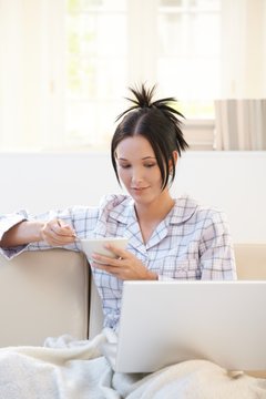 Young woman having breakfast in pyjama with laptop