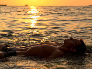 Muscle wet sexy naked man lying in sea water on sunset