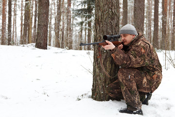 Man in camouflage aiming with sniper rifle at winter forest.