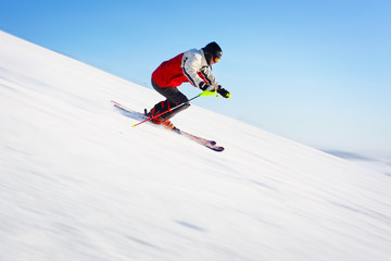 Skier in red jacket riding on hill. Motion blurred snow and clear blue sky