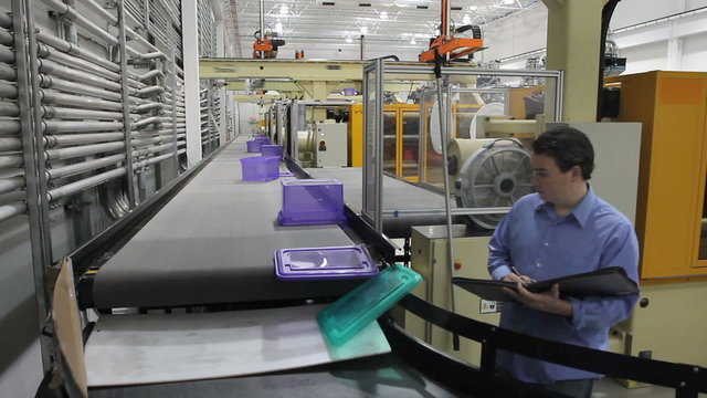A factory supervisor inspects products as they come down a conveyor belt with robotic arms moving in background.
