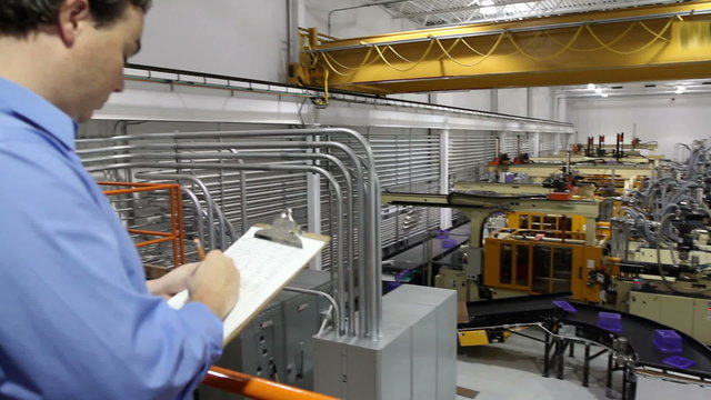 The camera pans across a large assembly factory and ends on a supervisor looking at his clipboard.

