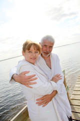 Senior couple in bathrobe standing on a pontoon - obrazy, fototapety, plakaty