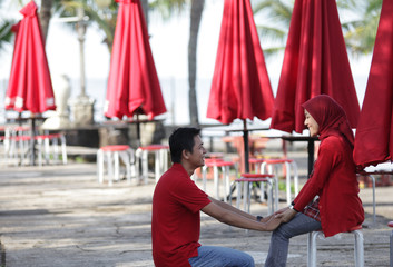 A cheerful Asian Muslim couple spending time together outdoors, dressed in matching red outfits. The man and woman are smiling and showing affection in a peaceful natural setting. This photo captures 