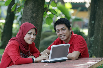 A cheerful Asian Muslim couple spending time together outdoors, dressed in matching red outfits. The man and woman are smiling and showing affection in a peaceful natural setting. This photo captures 