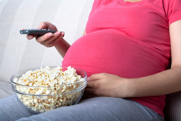 Pregnant woman sit and eat popcorn on sofa, watching television