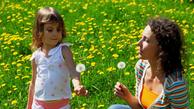 daughter playfully blows off dandelion in face of mother on lawn