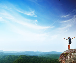 Young woman with a backpack standing on the cliff's edge with raised hands and looking to a sky