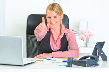 Smiling business woman sitting at desk and showing thumbs up