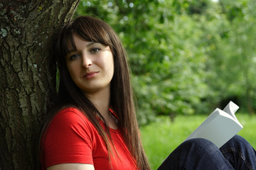 Young brunette girl in red shirt siting near tree and reading bo