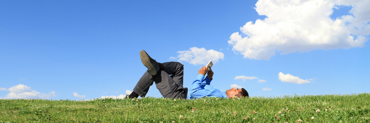 Businessman lying down on grass against blue sky with tablet. Man relaxing outdoors and working...