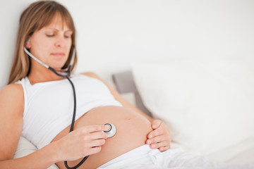 Beautiful pregnant woman using a stethoscope while lying on a be