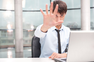 Fototapeta premium Angry young businessman sitting at his desk and showing stop ges
