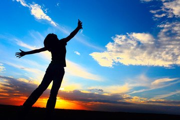 Fototapeta na wymiar Young woman standing on a meadow on sunset background and looking to a sky with raised hands.