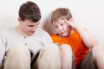 Two boys sit side by side with their backs to wall and read