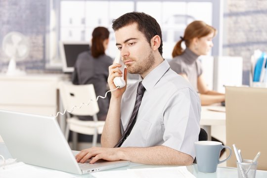 Young office worker using laptop talking on phone