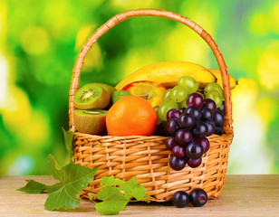 Ripe juicy fruits in basket on wooden table on green background