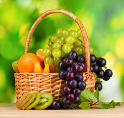 Ripe juicy fruits in basket on wooden table on green background