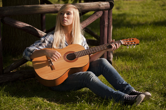 A young, beautiful blonde woman, dressed in a plaid shirt, leans against a rustic wooden fence while playing the guitar