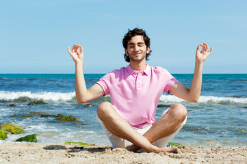 Fototapeta na wymiar Portrait of young man sitting in lotus pose on sand on beach and
