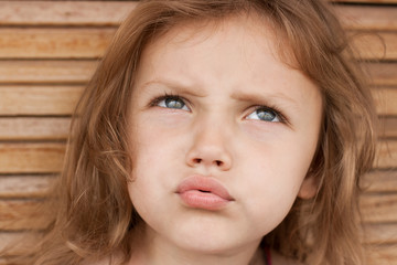 Closeup portrait of a young girl with a confused facial expression, thinking or pondering, outdoors against wooden background.