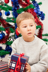 little boy sits with a gift near the dressed up New Year tree