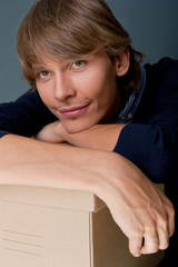 Portrait of young man leaning on box against grey wall. He is st