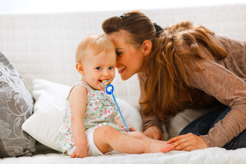 Adorable baby with soother and young mother playing on divan